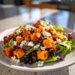 A close-up of a vibrant sweet potato salad featuring roasted orange sweet potato cubes, crumbled white cheese, and pecans over mixed greens.