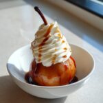 A beautifully presented poached pear topped with whipped cream and drizzled with caramel sauce, served in a white bowl.