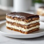 Close-up of a layered slice of chocolate eclair cake with creamy filling and dark chocolate ganache topping.