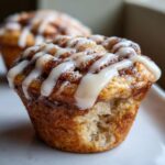 A close-up of a delicious cinnamon roll muffin with a bite taken out, drizzled heavily with white icing.
