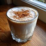 A glass of creamy coquito topped with a generous dusting of ground cinnamon, sitting on a wooden surface.