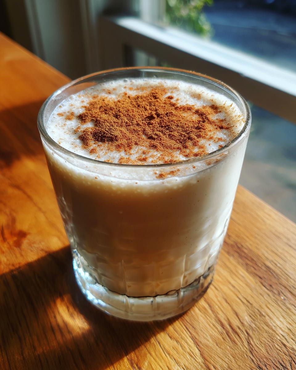 A glass of creamy, light-colored coquito topped generously with ground cinnamon, sitting on a wooden surface.