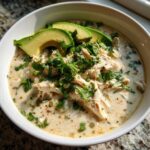 A close-up of a bowl of creamy keto white chicken chili topped with shredded chicken, fresh cilantro, and sliced avocado.