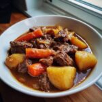 Close-up of a white bowl filled with rich crockpot beef stew, featuring tender beef chunks, bright carrots, and potatoes.