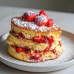 A tall stack of fluffy strawberry pancakes filled with macerated strawberries and topped with fresh berries and powdered sugar.