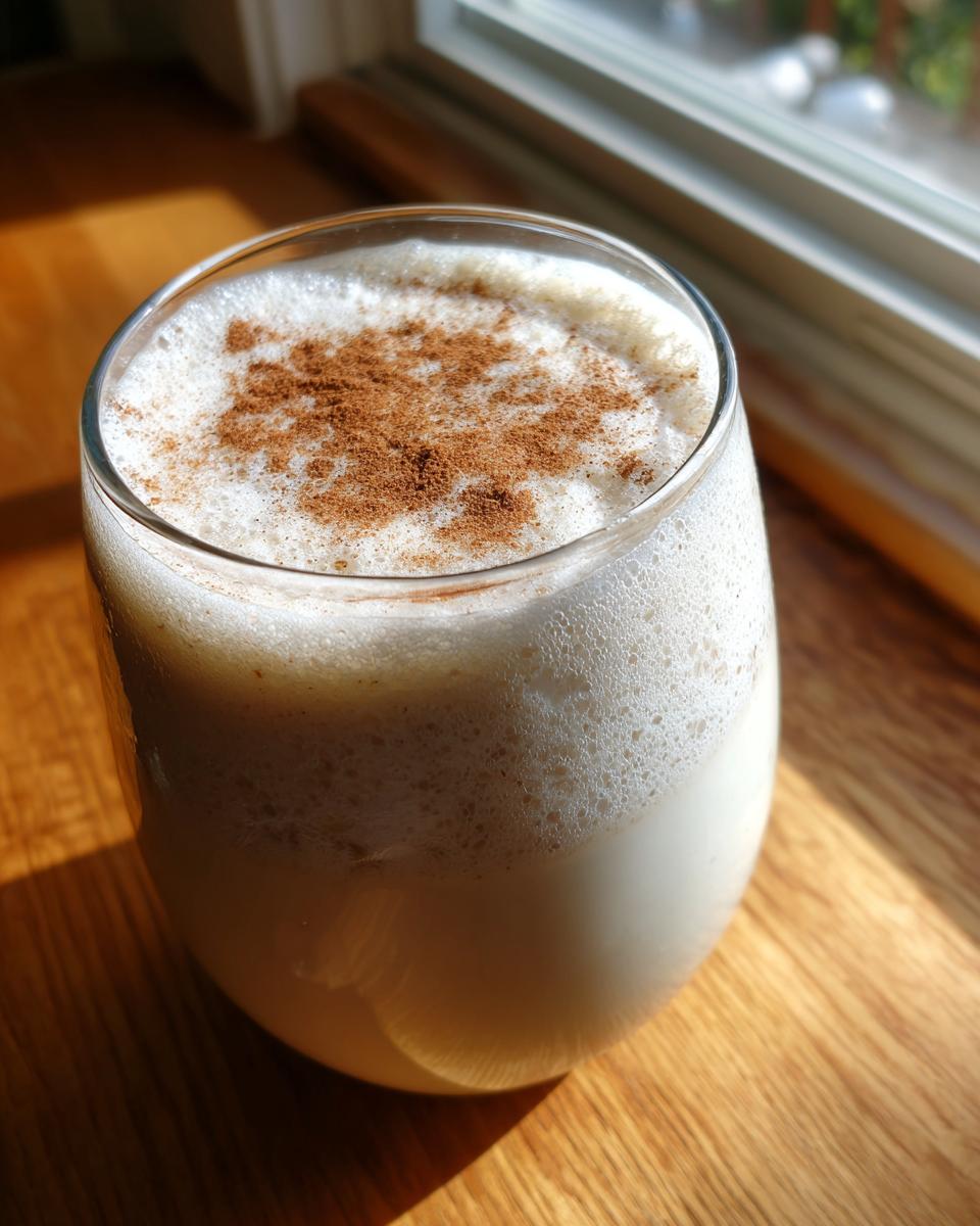 Close-up of a glass filled with creamy, frothy coquito topped with a sprinkle of ground cinnamon.