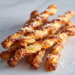A close-up of several golden brown, twisted cheese straws sprinkled with grated Parmesan cheese on a white marble surface.