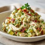A bowl filled with creamy honey mustard chicken salad mixed with avocado chunks, bacon bits, and fresh parsley.