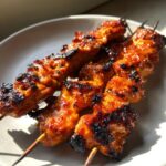 Three skewers of glossy, charred, and caramelized chicken skewers resting on a light plate.