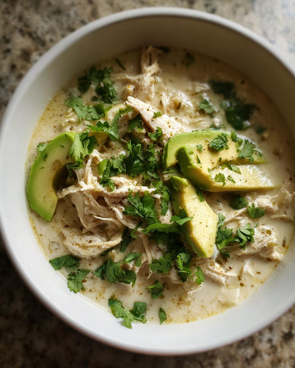 Close-up of a creamy bowl of keto white chicken chili topped with shredded chicken, fresh cilantro, and avocado slices.