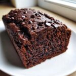A close-up of a rich, dark chocolate banana bread loaf topped with melted chocolate chips, resting on a white plate.