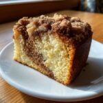 A close-up, sunlit slice of moist sour cream coffee cake featuring a yellow crumb and a thick cinnamon streusel topping.