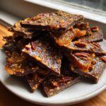 A stack of broken, glossy pieces of Pecan Pie Bark featuring dark chocolate and a crunchy pecan topping.