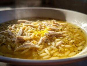 Close-up of a bowl of steaming penicillin soup featuring shredded chicken, orzo pasta, and bright yellow lemon zest.