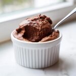 A close-up of rich, dark chocolate mousse served in a white fluted ramekin with a silver spoon resting in it.