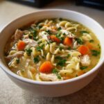 Close-up of a white bowl filled with savory turkey noodle soup, featuring shredded turkey, egg noodles, carrots, and parsley.