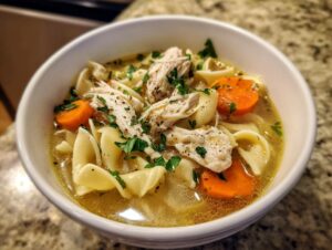 Close-up of a white bowl filled with savory turkey noodle soup, featuring shredded turkey, egg noodles, and sliced carrots.