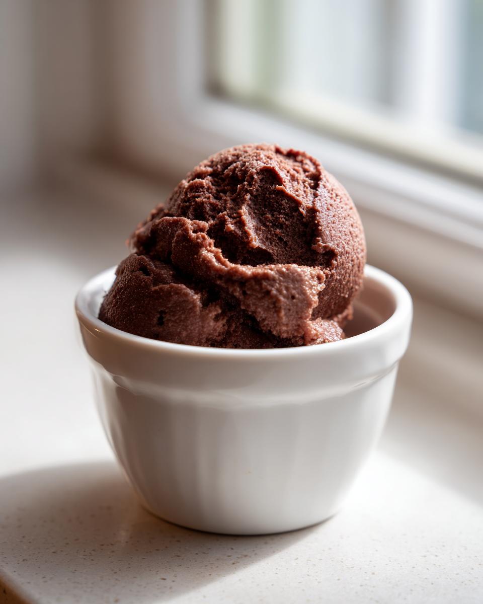 A single scoop of rich, dark chocolate mousse served in a small white ramekin, set near a window.