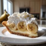A perfect slice of banana cream pie featuring a graham cracker crust, creamy filling, sliced bananas, and a generous topping of whipped cream.