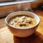 A steaming white bowl of creamy chicken chili topped with shredded chicken and grated cheese.