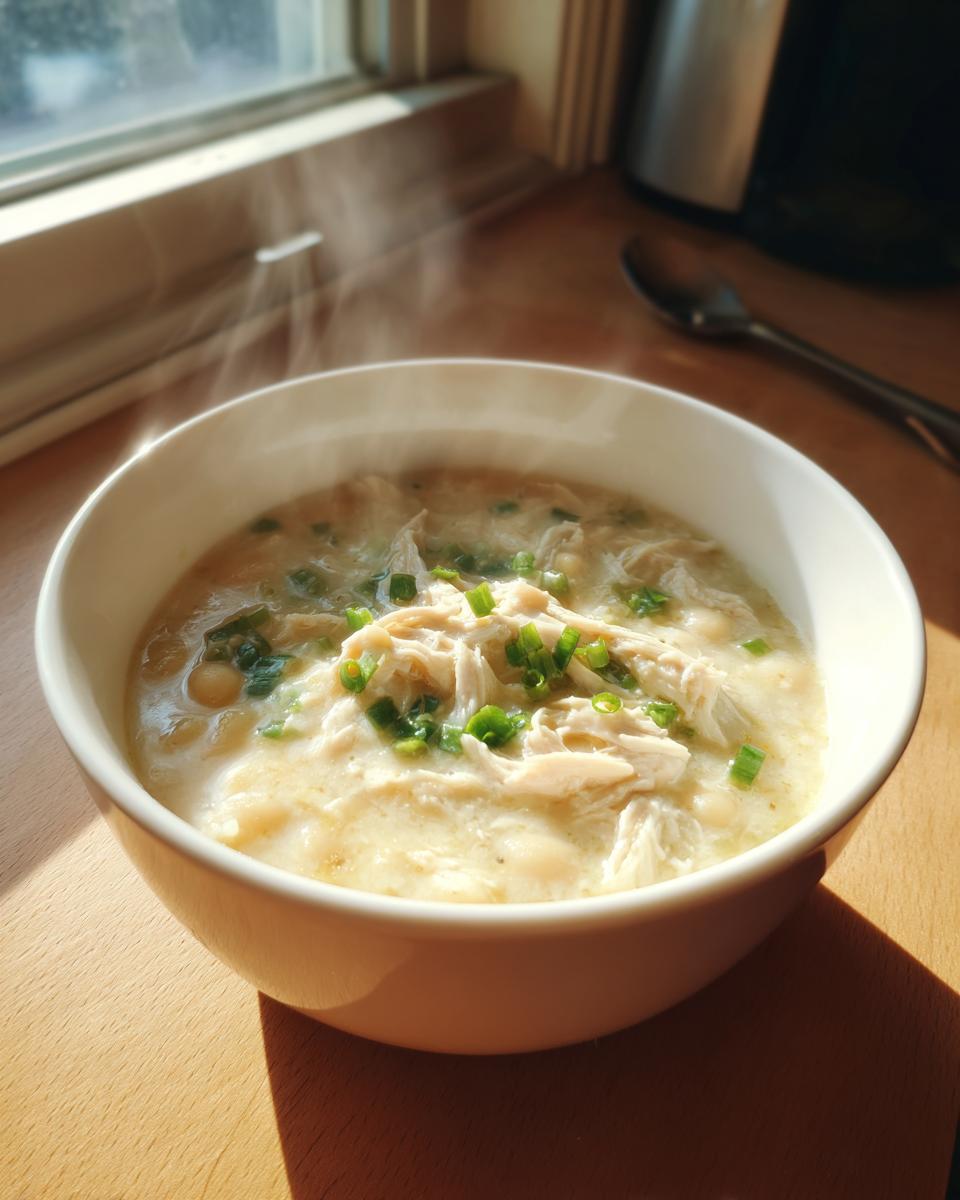 A steaming white bowl of creamy chicken chili topped with shredded chicken and fresh green chives.