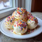A stack of white chocolate coated sugar cookie truffles generously topped with rainbow sprinkles on a white plate.